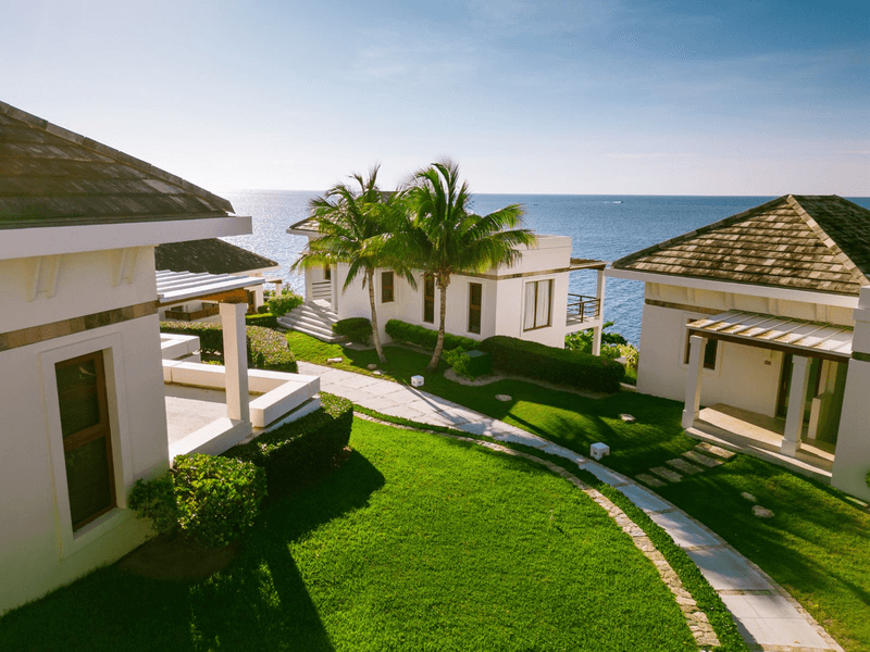Un moderno edificio de apartamentos o condominio de lujo de varios pisos con paredes exteriores blancas y grandes balcones. El piso superior cuenta con un área acristalada. Una larga piscina infinita es visible en uno de los balcones inferiores, rodeada de una terraza de madera y muebles de exterior. El edificio se encuentra en un entorno de exuberante bosque tropical verde.